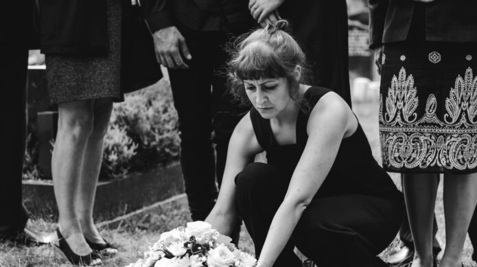 pouto-pribehy-lidi-po-smrti Černobílá fotografie ženy v černých šatech klečící u hrobu a pokládající smuteční kytici během pohřbu v kruhu rodiny.