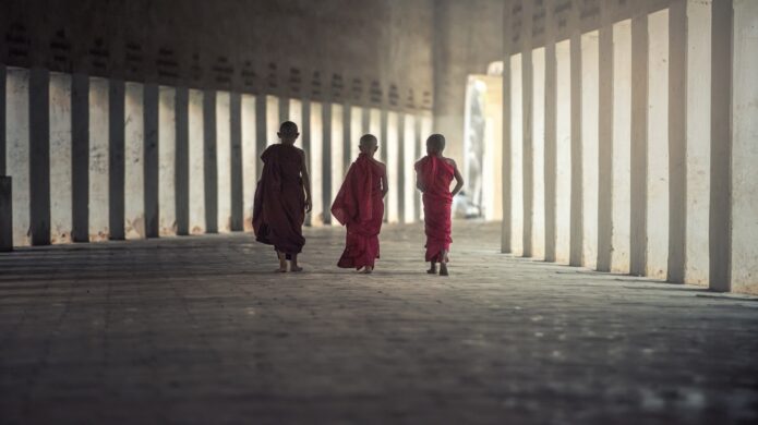 Mladí buddhističtí mniši kráčejí chodbou kláštera zalitou měkkým přirozeným světlem.